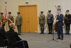 Przemawia minister spraw wewnętrznych i administracji Mariusz Błaszczak. W tle stoją przedstawiciele służb mundurowych. Z lewym rogu siedzą uczestnicy uroczystości.