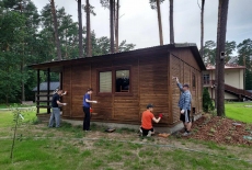 Na zielonej trawie stoi brązowy, drewniany dom, a przy nim czterech mężczyzn maluje ściany