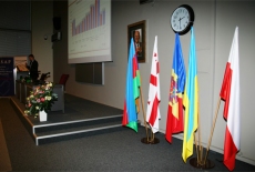 Na pierwszym planie widać flagi, a na drugim osobę stojącą przy mównicy.