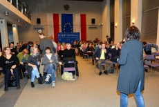 Zdjęcie ukazuje siedzących ludzi na widowni i Panią stojącą na scenie