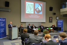 Aula KSAP, Bartosz Włodarski przemawia z mównicy, w prezydium siedzą pozostali paneliści. Na ekranie wyświetolny slajd przedstawiający symbol i odznakę Szkoły Nauk Politycznych UJ.