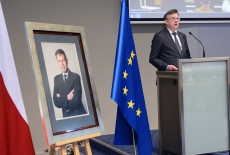 Szef Służby Cywilnej przemawia przy mównicy, obok stoi portret Władysława Stasiaka