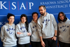 Pięć osób w szarych bluzach z napisem KSAP na tle niebieskiej ścianki reklamowej z logo KSAP.