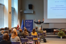 Uczestnicy konferencji w auli KSAP. Na mównicy Dyrektor KSAP. W podniesionych do góry rękach trzyma dwie książki.