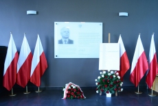 Tablica upamiętniająca Patrona KSAP. Wokół niej stoją polskie flagi, na podłodze leżą wiązanki biało-czerwonych kwiatów.