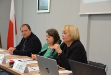 trzy osoby siedzą przy ławce, jedna z nich mówi przez mikrofon