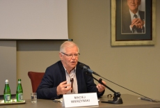 Maciej Wierzyński siedzi przy stole prezydialnym i prowadzi wykład.