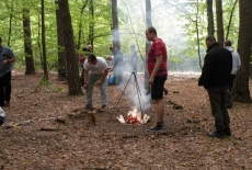 Dwóch słuchaczy rozpala ognisko w lesie, a wokół nich pozostali uczestnicy wyprawy.
