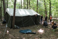 Sześć osób rozbija duży, zielony, wojskowy namiot w lesie