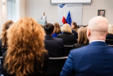 Uczestnicy siedzą w auli. Prowadzący stoi przy mównicy.