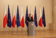 Tomasz Arabski stoi przy mównicy i przemawia na tle flag Polski i UE