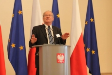 Pan przemawia na mównicy na tle flag Polski i UE
