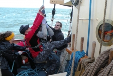 Czterech mężczyzn ciągnie liny ożaglowania w czasie żeglugi.