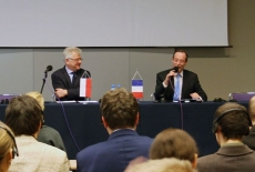 Ambasador Francji siedzi przy stole prezydialnym i mówi do mikrofonu, obok siedzi dyrektor KSAP