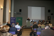 Dyrektor KSAP Jacek Czaputowicz przemawia na mównicy do zgromadzonych uczestników. Obok przy stole siedzą prowadzący