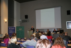 Dyrektor KSAP Jacek Czaputowicz przemawia na mównicy do zgromadzonych uczestników. Obok przy stole siedzą prowadzący