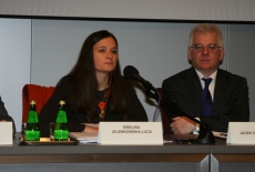 Pani Ewelina Jelenkowska-Luca siedzi przy stole i mówi do mikrofonu. Obok siedzi dyrektor KSAP