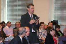 Uczestnik konferencji stoi i mówi do mikrofonu obok siedza inni uczestnicy