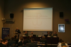 Dyrektor KSAP Jacek Czaputowicz przemawia na mównicy, obok przy stole prezydialnym siedza prowadzący konferencje