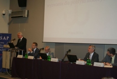 Dyrektor KSAP Jacek Czaputowicz przemawia na mównicy, obok przy stole prezydialnym siedza prowadzący konferencje