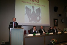 Wojciech Kalwat przemawia na mównicy obok za stołem prezydialnym siedzą Piotr Szlanta, Jan Pastwa, Piotr Koryś, Jakub Skiba