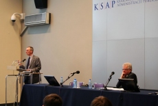 prof. Christopher Pollit stoi i przemawia przy mównicy obok przy stole prezydialnym siedzi Dyrektor KSAP