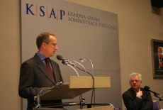 Pan Jan Tombiński stoi przy mównicy i przemawia obok przy stole siedzi Dyrektor KSAP