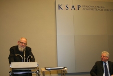 prof. Bohdan Cywiński stoi przy mównicy i przemawia, obok siedzi Dyrektor KSAP
