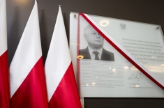 Tablica pamiątkowa przedsawiająca portret Prezydenta Lecha Kaczyńskiego oraz nadrukowany tekst. Obok tablicy stoją trzy flagi biało-czerwone.