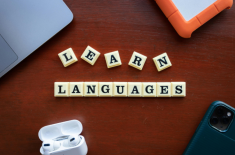 Klocki z literami układające napis „LEARN LANGUAGES” na drewnianym biurku. Wokół widoczne są laptop, smartfon, słuchawki bezprzewodowe oraz dysk zewnętrzny.