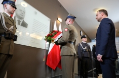 Prezydent Andrzej Duda, w asyście wojskowej, składa kwiaty pod tablicą pamiątkową. 