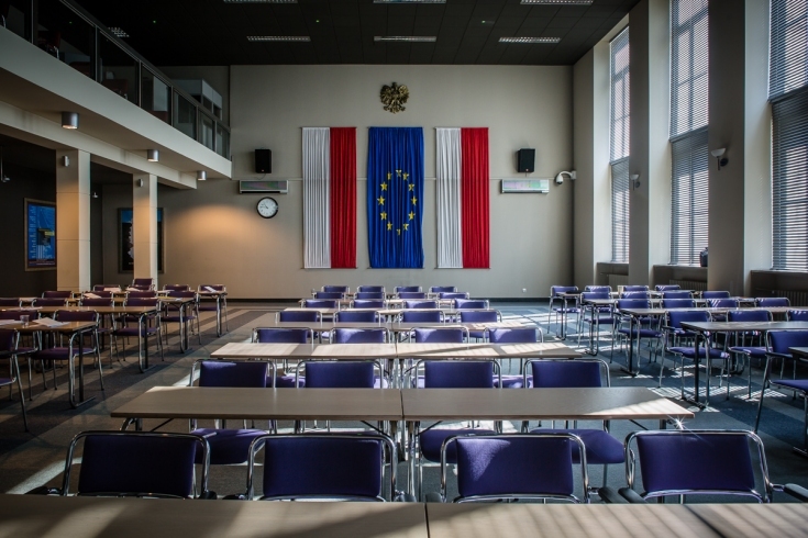 Widok na aule KSAP. Po środku stoją stołu z krzesłami, na ścianie wiszą dwie flag Polski a po środku flaga UE.