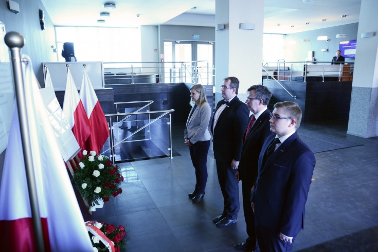 przedstawaiciele KSAP oraz Szef Służby Cywilnej stoją przed tablicą upamiętniającą Patrona KSAP. Widoczne polskie flagi oraz wiązanki.