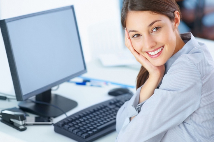 Uśmiechnięta kobieta siedzi przy biurku, na którym znajduje się monitor i klawiatura od komputera.