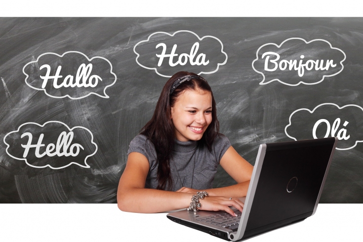 dziewczyna siedzi przed otwartym laptopem, w tle na czarnej tablicy napisy w różnych językach
