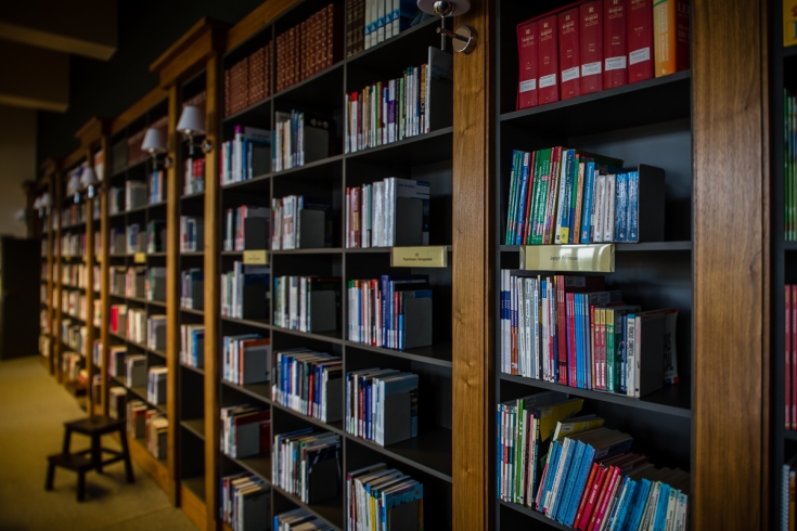 Regał w bibliotece na którym stoją książki. Obok stoi stołek.